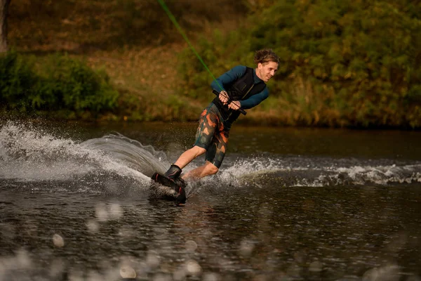 Allegro giovane uomo in muta e gilet cavalca wakeboard sul fiume — Foto Stock