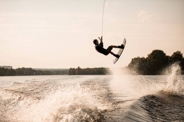 İp tutan enerjik kaslı adamın görüntüsü ve ustaca su sıçratma üzerine wakeboard ile taklalar atıyor.