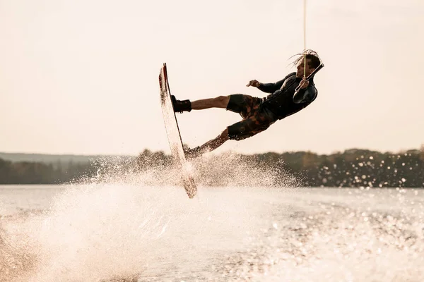 Energetic muscular man holds rope and makes flips with wakeboard over splashing water — Stock Photo, Image