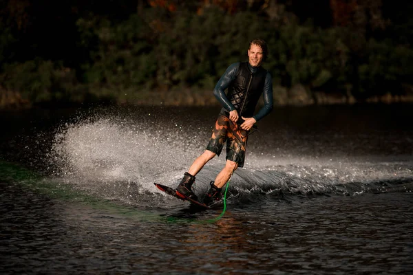 Felice giovane uomo in giubbotto di salvataggio nero tiene corda verde brillante e cavalca wakeboard — Foto Stock