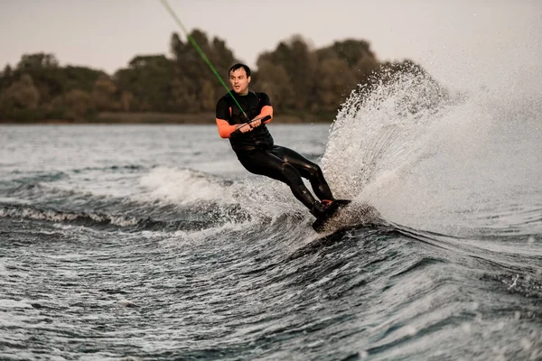 Volwassen man in wetsuit houdt kabel in zijn handen en wakeboarden op spetterende golf — Stockfoto