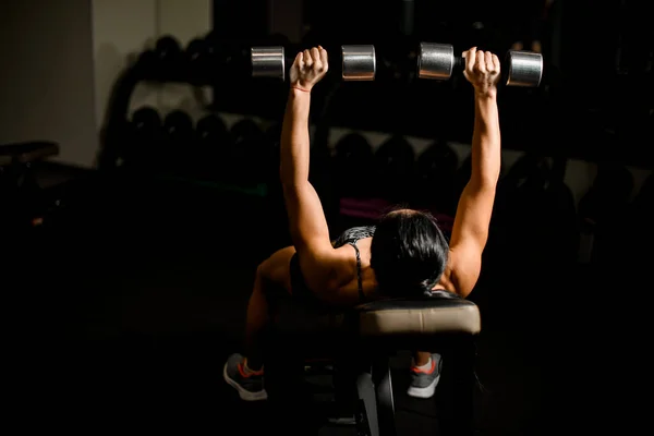 Atletisk brunett kvinna träning med hantlar liggande på bänk i gym. — Stockfoto
