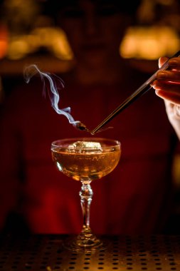 Barın üstünde buzlu bir içecek ve barmen yanan bir bitki tutuyor.