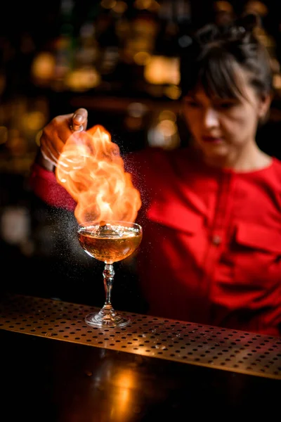 Crystal glass with iced drink stands on bar and barman sprinkles and sets fire over it — Stock Photo, Image
