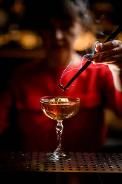 Beautiful crystal glass with iced drink on bar and bartender holds smoldering plant over it — Stock Photo, Image