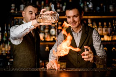 Barmen bardağa su doldurur ve başka bir barmen bardağı ateşe verir.