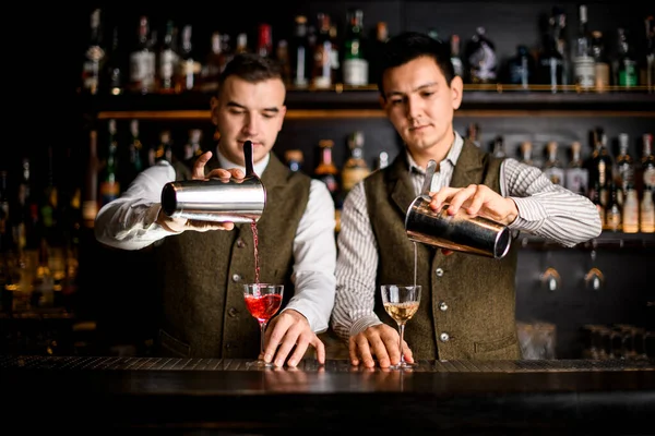 two bartenders pour cocktails into glasses at the same time at bar