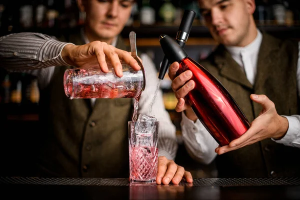 Vista de vidro em que dois bartenders simultaneamente derramar líquido de sifão e xícara — Fotografia de Stock