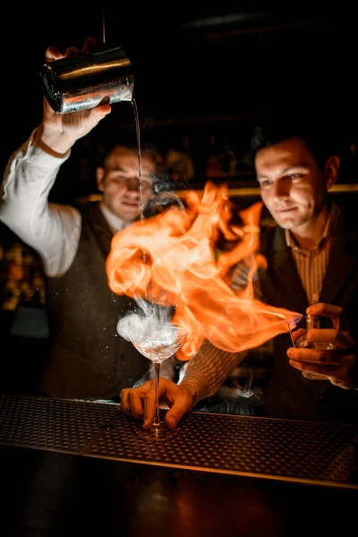 Barman derrama líquido em vidro de bebida fumegante e outro homem polvilha sobre ele e incendeia — Fotografia de Stock