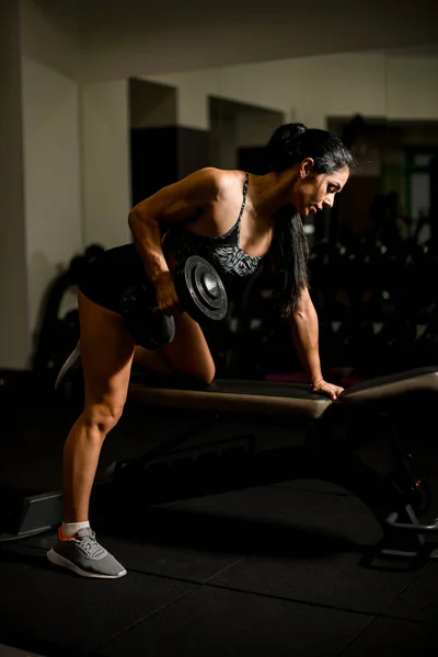 Sidovy av kvinnlig kroppsbyggare som tränar i gym med hantel i handen på simulator. — Stockfoto