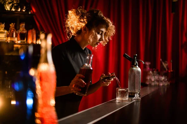 Νεαρή γυναίκα μπάρμαν στέκεται πίσω από το μπαρ και χύνει ποτό από jigger σε γυαλί — Φωτογραφία Αρχείου