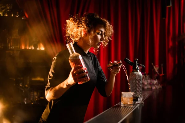 Ελκυστική γυναίκα μπάρμαν κρατώντας ένα μπουκάλι αλκοόλ στο χέρι της και χύνει ποτό από jigger — Φωτογραφία Αρχείου