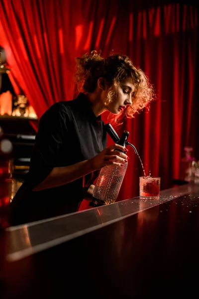 Woman bartender stands behind the bar and pours drink from siphon into glass — Stock Photo, Image