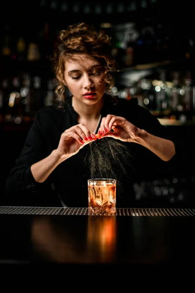 Beautiful female bartender gently sprinkles on glass with cold alcoholic cocktail on the bar counter — Stock Photo, Image