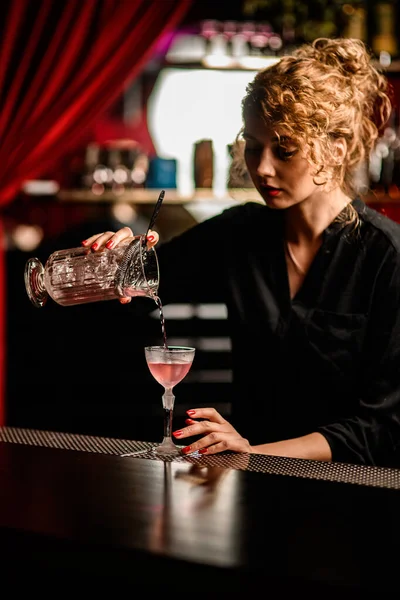 View of beautiful female bartender pouring cocktail from mixing cup into crystal glass — Stock Photo, Image