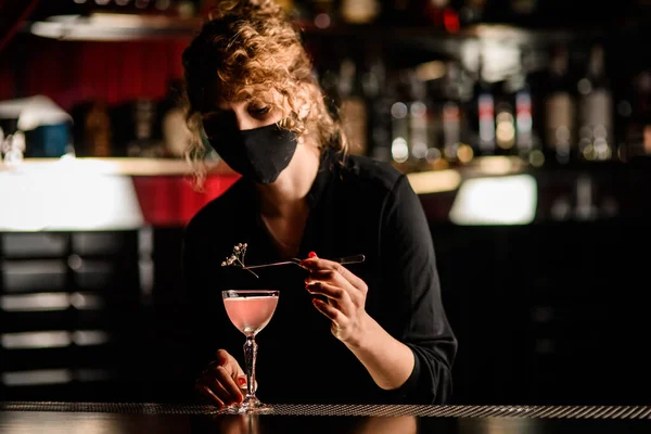 Вид барменши в маске, аккуратно украшающей стакан коктейля цветочной веточкой — стоковое фото