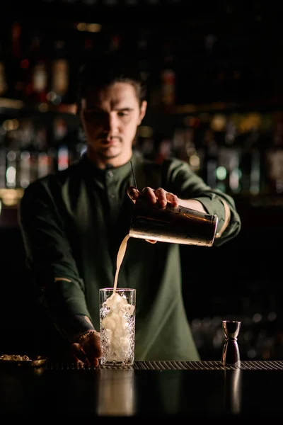 Jonge barman schenkt cocktail van stalen shaker in glas in donkere bar — Stockfoto