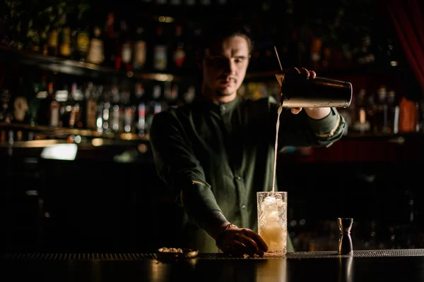 Uitzicht op jonge barman gieten cocktail van stalen shaker in glas met ijs — Stockfoto