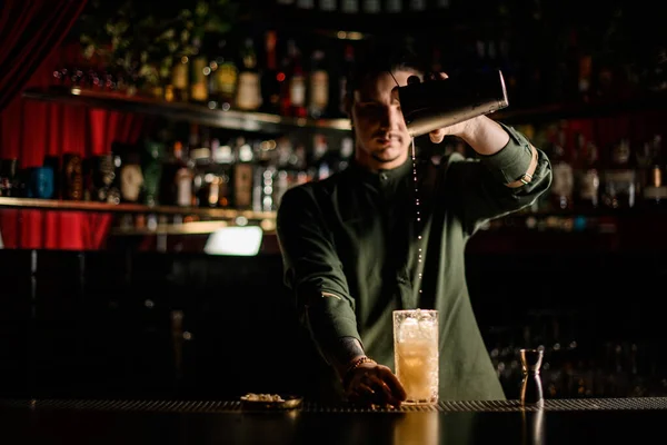 Uitzicht op jonge barman gieten cocktail van stalen shaker in glas in donkere bar — Stockfoto