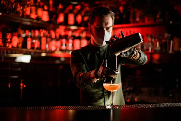 Profi-Barkeeper in Maske gießt Cocktail aus Shaker durch Sieb sanft ins Glas — Stockfoto