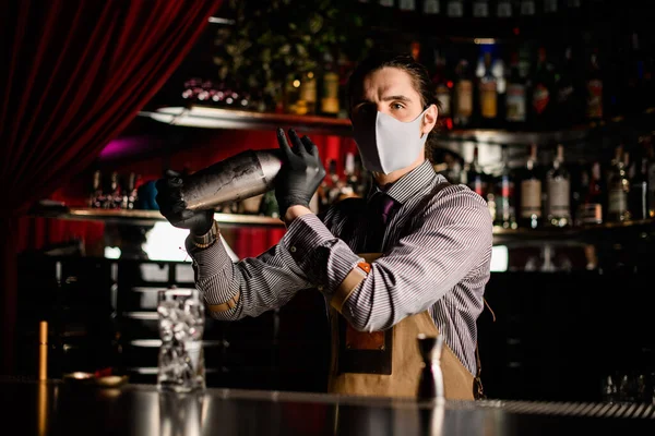 Barman in schort en masker en handschoenen staat achter de bar en houdt stalen shaker — Stockfoto