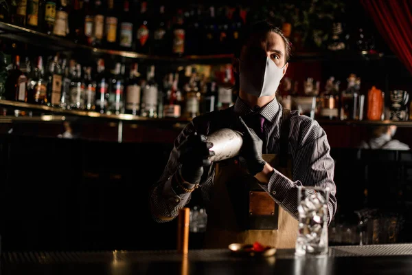 Bartender i förkläde och mask och handskar står bakom baren med stål shaker — Stockfoto