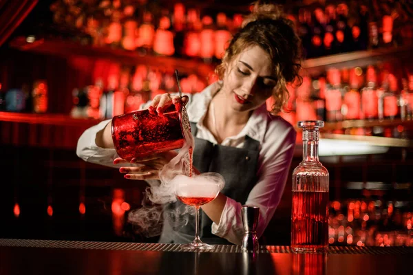 Jovem barman feminino derramando coquetel fumegante brilhante em vidro no balcão de bar — Fotografia de Stock