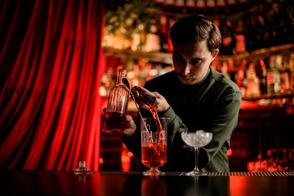 Joven hombre cuidadosamente verter bebidas alcohólicas brillantes en la taza de mezcla en el mostrador de bar — Foto de Stock