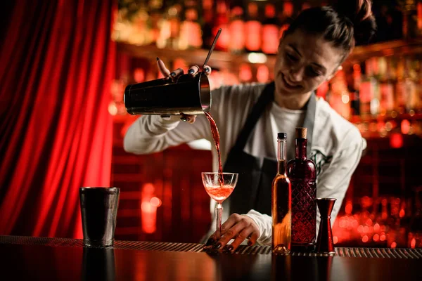 Glimlachende vrouw barman zachtjes schenkt cocktail van shaker in glas aan bar teller — Stockfoto