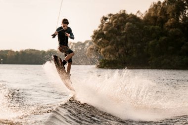 Genç erkek wakeboardcu sıçrama dalgasında gemiyle atlıyor.