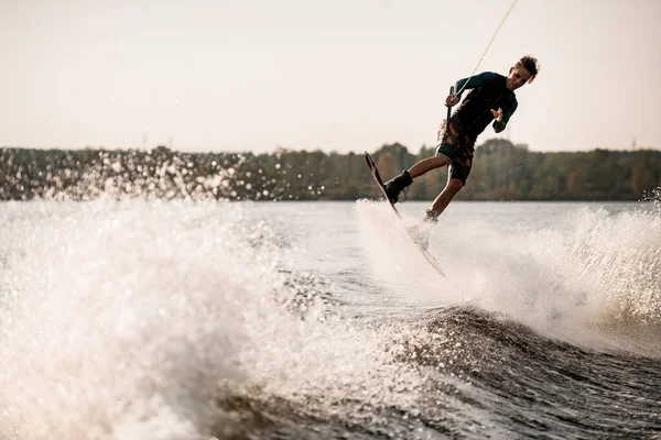 Young man wakeboarder holds cable and actively rides on splashing wave. — Stock Photo, Image