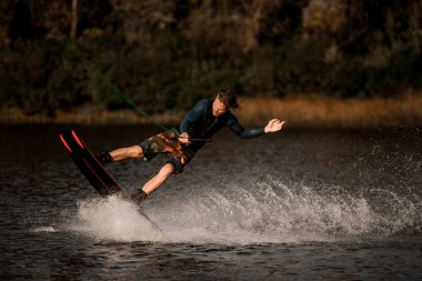 Dalgıç kıyafetli yakışıklı adam gergin bir ip tutuyor ve su yüzeyinde wakeboard sürüyor.