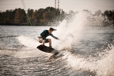 Can yeleği giymiş erkek wakeboardcu elleriyle ipi tutuyor ve su sıçratan dalgalarda dengeyi sağlıyor.