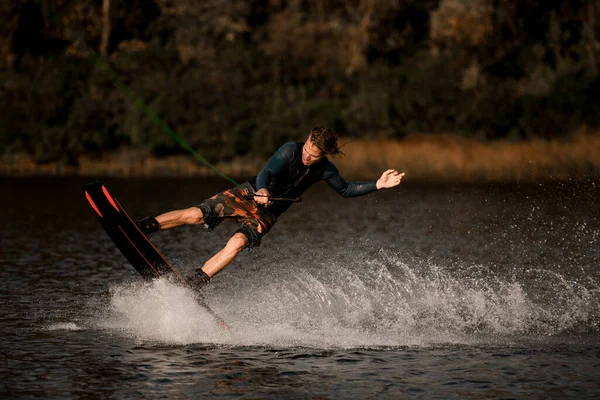 Hombre deportivo guapo en traje de neopreno sostiene cuerda tensa y monta wakeboard en la superficie del agua —  Fotos de Stock