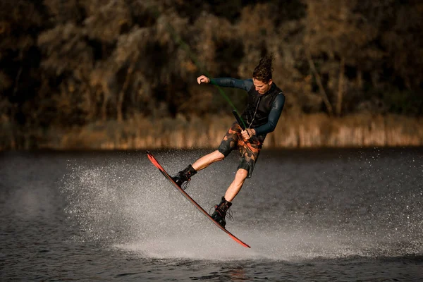 Vista di atletico ragazzo muscolare salta attivamente sul wakeboard tenendo la corda in mano. — Foto Stock