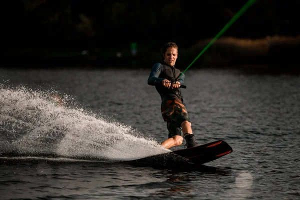 Uomo atletico in muta magistralmente in equilibrio e cavalcando sul wakeboard veloce su spruzzi d'acqua di fiume — Foto Stock