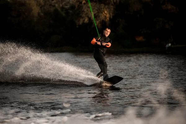 Uomo atletico in muta cavalca energicamente sull'onda del fiume spruzzata sul wakeboard. — Foto Stock