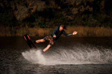 Dalgıç kıyafetli sporcu gergin bir ip tutuyor ve su yüzeyinde wakeboard sürüyor.