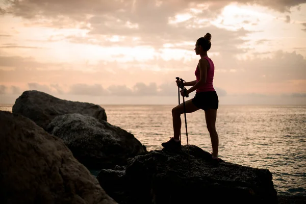 Kvinna med trekking pinnar står på sten mot bakgrund av hav och himmel — Stockfoto