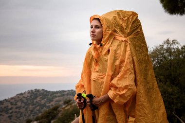 Sırt çantalı, sarı yağmurluklu bir kadın gün doğumuna karşı ayakta duruyor..