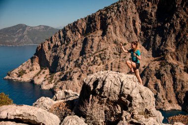 Yürüyüş sopalı bir kadın dağların ve deniz koyunun arka planına karşı büyük bir kayaya tırmanır..