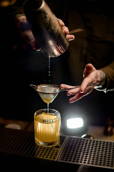man at bar gently pours cocktail from shaker cup into old-fashioned glass through sieve