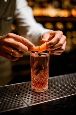 Barmenlerin ellerinin portakal kabuğuyla süslediği soğuk bir içecekle kristal bardağa bak.
