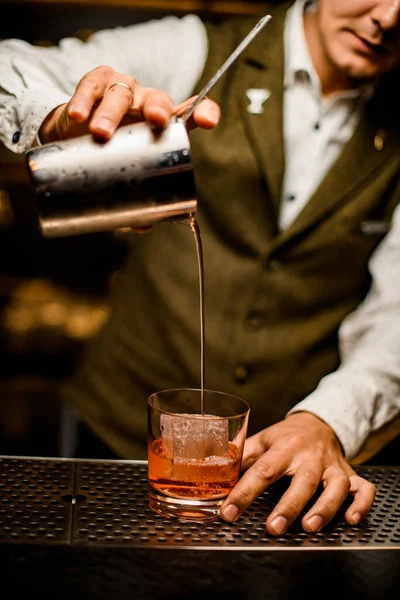 man at bar professionally pours drink from steel mixing cup into old-fashioned glass