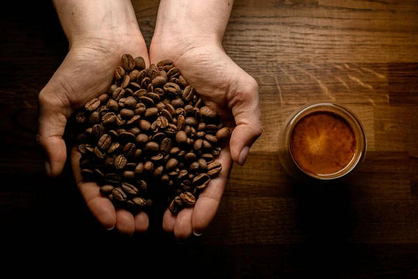 Hænder med ristede kaffebønner over træoverflade og kop kaffe i nærheden - Stock-foto