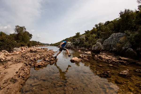 Чоловічий турист з пішохідними полюсами стрибає на каменях через річку — стокове фото