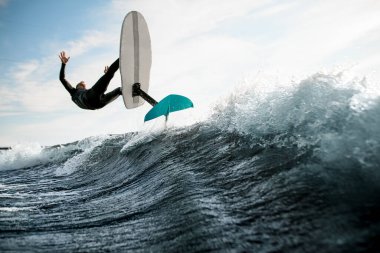 Siyah dalgıç kıyafetli bir adam, elinde folyoyla dalganın üzerinden atlıyor.