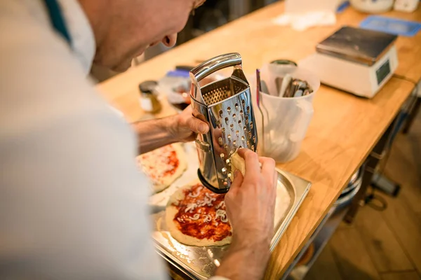 Mężczyzna szef kuchni trzyma metalową tarkę i tarki ser na bazie pizzy — Zdjęcie stockowe