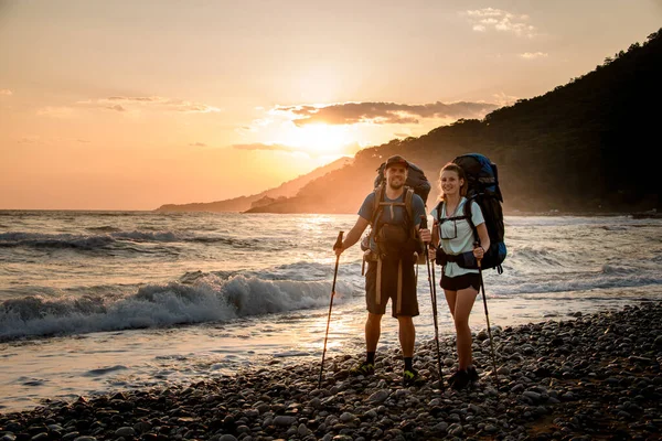 Feliz joven hombre y mujer turistas en la orilla del mar contra el telón de fondo de la puesta del sol —  Fotos de Stock