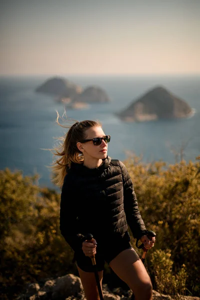 Vista de la hermosa mujer con gafas de sol y bastones de senderismo en el fondo del paisaje marino — Foto de Stock